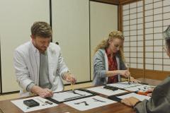 Two students and an instructor participate in a calligraphy class at a long wooden table in front of a traditional Japanese shoji screen.