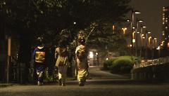Three women in traditional Japanese attire walk down a street at night, illuminated by streetlights and the distant glow of city buildings.
