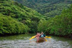 A colorful sabani boat with four people is paddling down a calm river surrounded by lush green trees and hills.