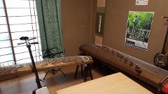 Dette billede viser et traditionelt japansk rum med musikinstrumenter såsom en shamisen og to kotoer, sammen med traditionel indretning som et vindue med shoji-skærme og en hængende skriftrulle.