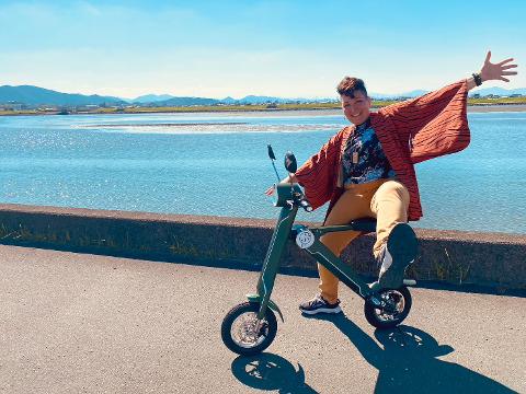 Ise Day Tour: Explore by Electric Tuk-Tuk or Bike with Guide A person on an electric scooter with their arm outstretched, posing in front of a body of water.