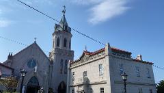 A low-angle shot shows a majestic grey stone church with a towering steeple topped by a weather vane, connected to a building with a red roof, against a clear blue sky.