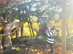 Un couple se tient en prière devant un petit sanctuaire niché sous un grand arbre soutenu par des supports, sur les rives de la rivière Shiodomari.