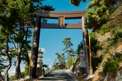 A large brown torii gate stands before a road leading up a hill, surrounded by lush green trees and a clear blue sky.
