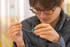 A person with glasses meticulously crafts a traditional Japanese mizuhiki knot with white and green cords.