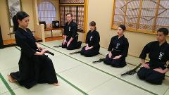 Five individuals in black hakama and gi kneel on tatami mats, each with a sword in front of them, in a traditional Japanese dojo.