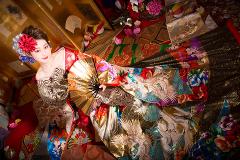 Eine Frau in einem farbenfrohen Kimono und mit geblümtem Haarteil hält einen goldenen Fächer, umgeben von kunstvollen Stoffen und Dekorationsgegenständen in einer prächtigen, traditionellen japanischen Umgebung.