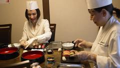 Two women wearing chef hats and gloves are making sushi together at a wooden table with various ingredients and tools.