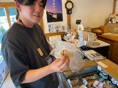 A young man holds up a wrapped pastry in front of a shop counter filled with goods and a cash register.