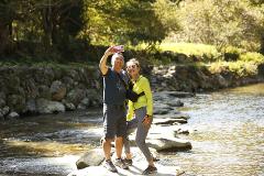 A couple poses for a selfie while standing on rocks in a river.