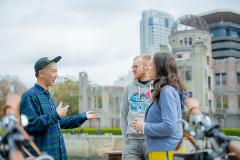 Two tourists listen to a Japanese man talk in front of the Hiroshima Peace Memorial, bicycles are visible in the foreground.