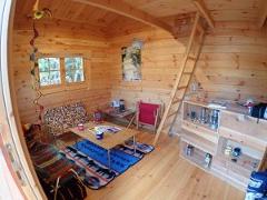 The image shows the cozy interior of a wooden cabin with a ladder, seating, a table, and shelves filled with camping gear, suggesting a comfortable resting spot after outdoor activities.