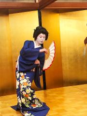 一名日本艺伎身穿带有花卉图案的蓝色和服，脸上涂着白色颜料，手持一把红边白扇，在金色屏风前表演。