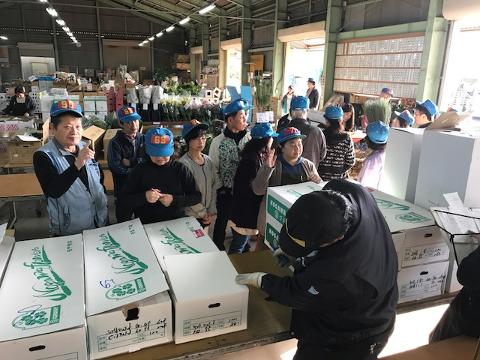 A group of people wearing blue hats with numbers are gathered around boxes labeled "Nogawa Flowers" in a warehouse-like setting.