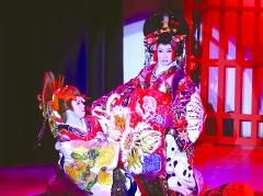 Two Kabuki actors in elaborate costumes and makeup, one seated and one standing, perform on a stage with red lighting.