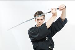 A young man in a black gi holds a katana sword above his head against a white background in this image promoting Iaido in Asakusa.