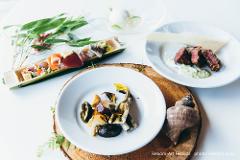 A beautiful Japanese meal consisting of sashimi in bamboo, grilled meats with avocado dip, and a dish of mushrooms and vegetables is artfully arranged on a white tabletop with bamboo leaves and a seashell.