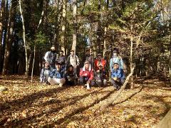 A group of people in outdoor clothing pose for a photo in a forest floor covered in fallen autumn leaves.