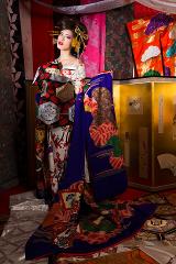 Eine Frau im Kimono steht umgeben von bunten Stoffen und einem goldenen Paravent.