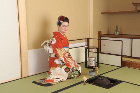 Kirkkaanpunaiseen kimonoon pukeutunut nainen poseeraa perinteisessä japanilaisessa teehuoneessa istuen tatami-matoilla teeseremonian läheisyydessä.
