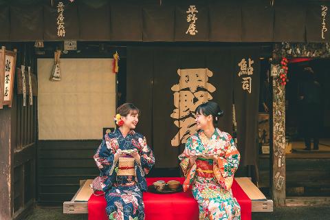 Två kvinnor i färgglada kimonos sitter och ler mot varandra utanför en traditionell japansk butik, vilket skapar en varm och inbjudande miljö.