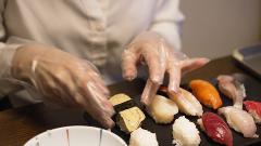 A person wearing plastic gloves is preparing a variety of sushi on a black plate, including tuna, salmon, shrimp, and egg sushi.
