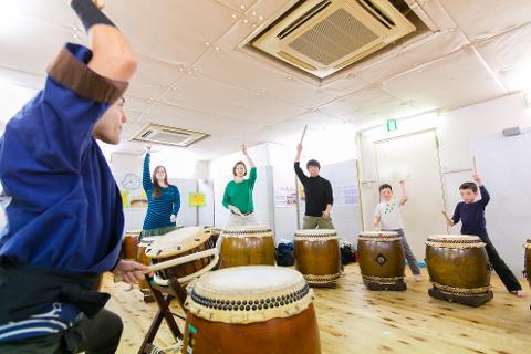 Um grupo de pessoas, incluindo adultos e crianças, está aprendendo a tocar tambores taiko japoneses em uma sala iluminada com um professor em primeiro plano.