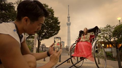 A man takes a photo of a woman in a rickshaw, with the Tokyo Skytree in the background.