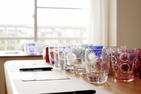 A row of colorful, ornate Edo Kiriko glasses sits on a wooden table in front of a window.