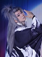 A person with long gray hair in a high ponytail, wearing dramatic makeup and a black and white robe, poses with their hand near their face.