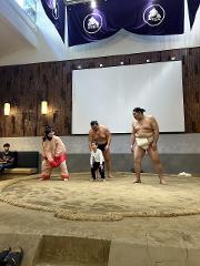 A sumo show takes place in a restaurant with a sumo ring, where two sumo wrestlers and a child are present.