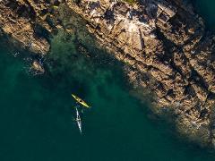 An aerial view captures two kayakers navigating clear waters near a rocky coastline with a captivating contrast of textures and colors.