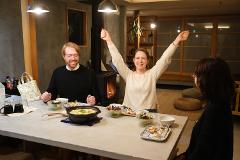 Two people are seated at a table with food, one cheering with arms raised, while another person sits with their back to the camera.