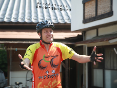 A man in a cycling outfit and helmet gestures with his hands, standing in front of a traditional Japanese building with a tiled roof.