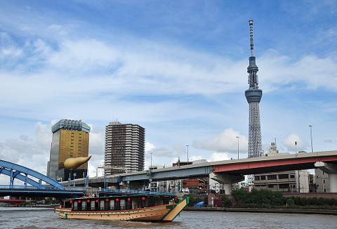A traditional Japanese boat sails on a river with a modern city skyline featuring the Tokyo Skytree and other skyscrapers in the background under a cloudy sky.