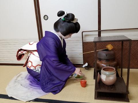 A geisha in a purple kimono kneels on a tatami mat, preparing a matcha tea ceremony.