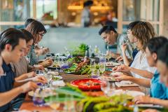 A group of people are seated at a long table, enjoying a meal with drinks, adorned with floral decorations in a bright, airy setting.