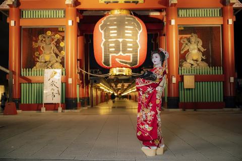 A geisha in a red kimono stands before a large red lantern in front of a Japanese temple at night.