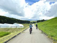 Two people ride e-bikes down a paved path through a landscape featuring greenhouses, green fields, and rolling hills under a cloudy sky.