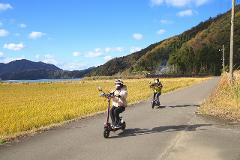 Two people on electric scooters ride along a road beside a golden field with a lake and mountains in the background.