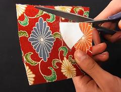 A close-up shot shows hands cutting a square piece of red patterned fabric with scissors, part of a craft activity.