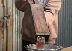 A person in a tan sweatshirt holds a wooden mold to extract a clear amber-colored substance into a metal sieve.