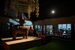 A sculpture of a Buddha atop an elephant is displayed under a canopy of golden bells, with people watching a taiko drum performance in the background.
