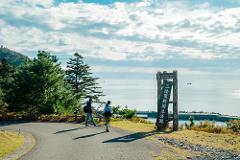 Two figures walk on a path towards the sea with a national park sign in Sanriku Fukko National Park.