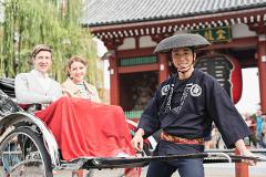 A rickshaw driver in a traditional hat and uniform poses in front of a temple with a couple seated in the rickshaw.