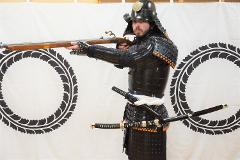 A person in full samurai armor with a musket and two swords stands before a white backdrop with circular designs.