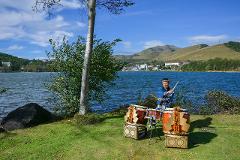 A man in traditional Japanese attire plays taiko drums by Lake Shirakaba, with rolling hills and a town in the background.