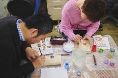 Two people are practicing the Japanese art of Kiri wood inlay crafting, meticulously painting intricate designs onto wooden pieces with brushes.
