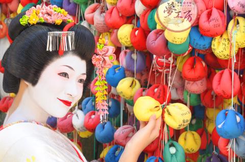 A woman dressed as a Japanese Geisha stands in front of a wall of colorful prayer pouches, touching a yellow one.