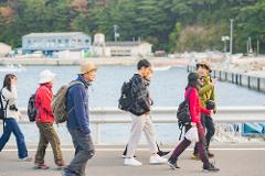 A group of tourists in casual wear with backpacks walk along a waterfront path with trees in the background.